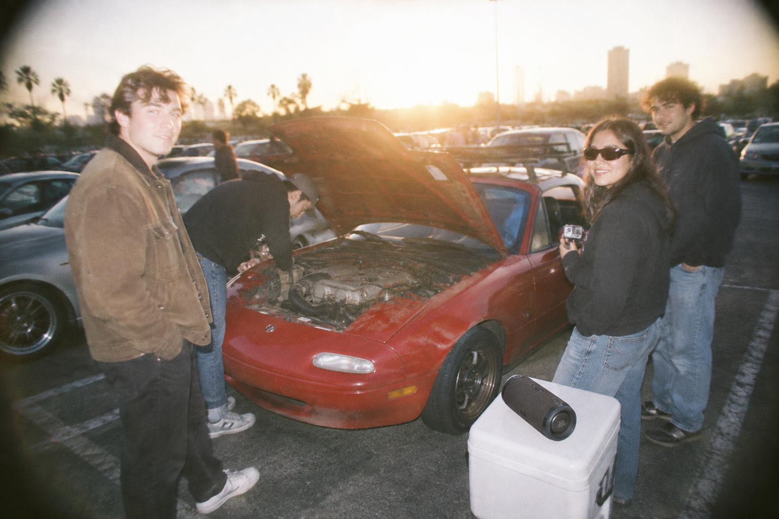 Four friends at a sunset parking-lot car meet around a red Miata with its hood up.
